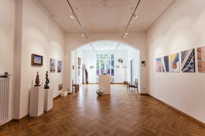 Lege galeriezaal bij NUMMER18: schilderijen en sculpturen op sokkels links, grote abstracte fotowerken rechts, met een lage boog die uitkomt op een tweede zaal met daglicht