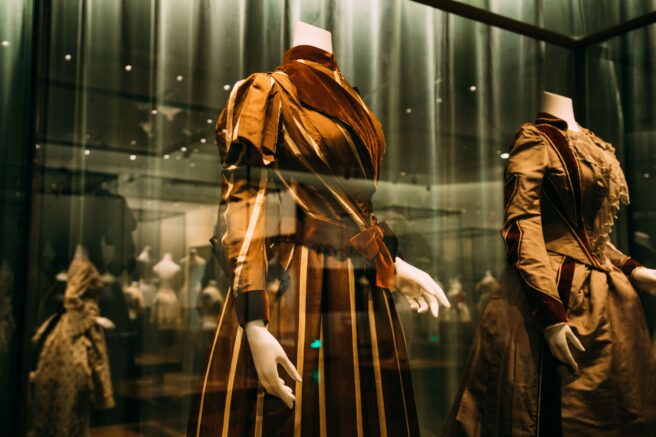 Twee historische japonnen op hoofdloze mannequins achter museumglas, warm verlicht in goudtinten — sfeerbeeld bij speeddaten in Modemuseum Hasselt