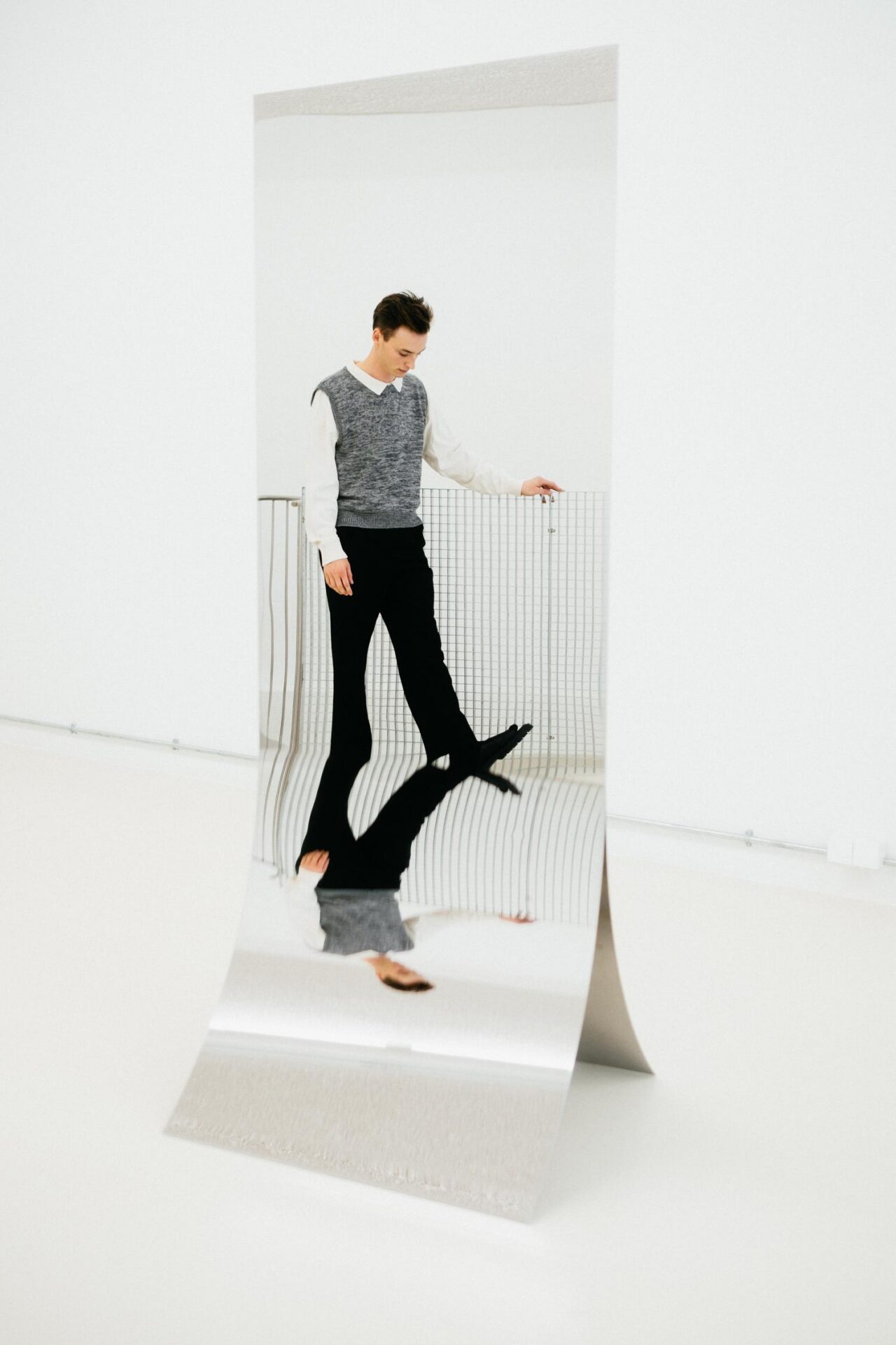Een hoge, gebogen plaat gepolijst aluminium staat op de vloer van een witte expositieruimte en weerspiegelt een persoon in een grijze spencer en een zwarte broek, met gebogen hoofd; onder de knik in de plaat staat het spiegelbeeld omgekeerd.