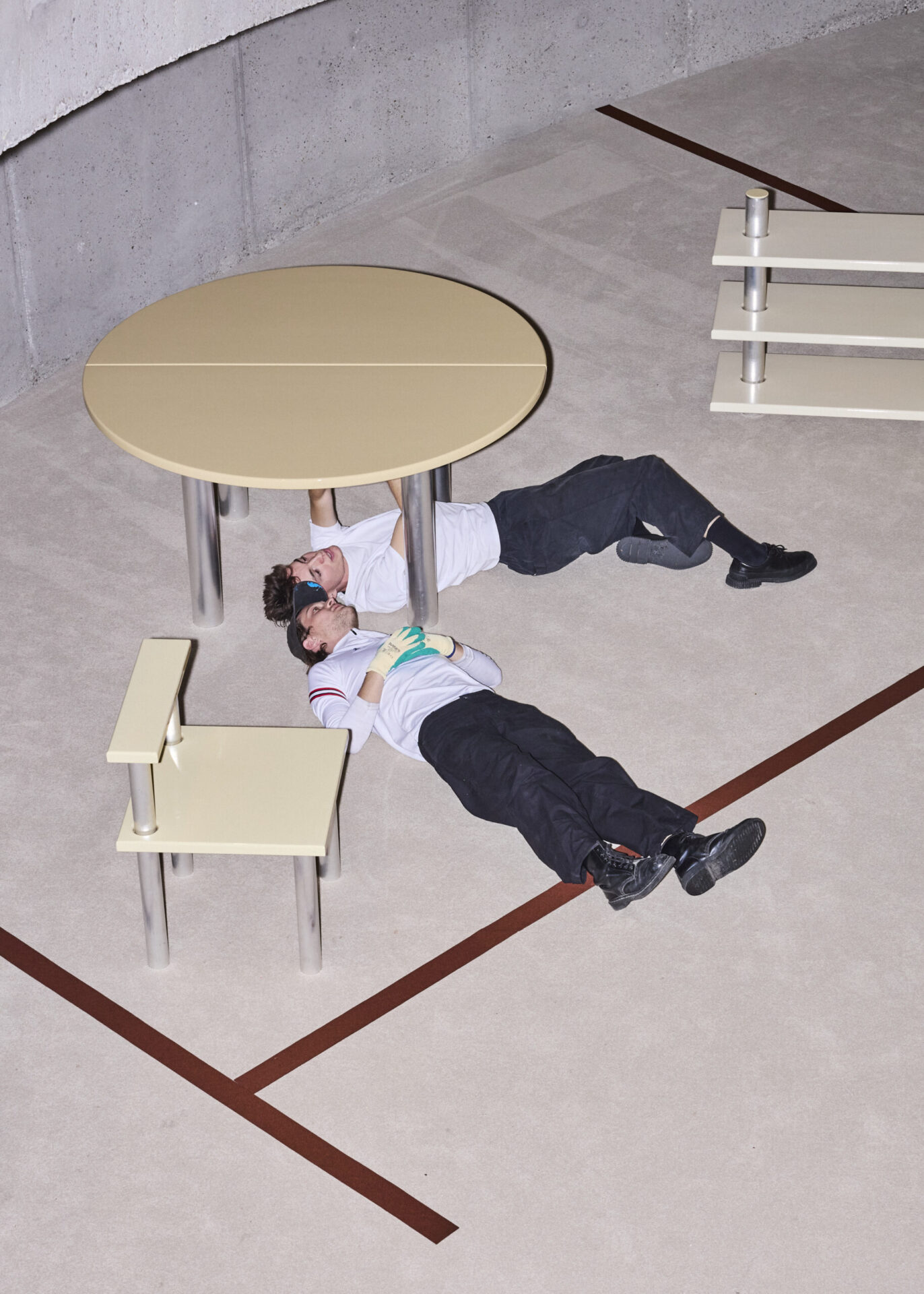 Twee personen in witte polo's en donkere broeken liggen op een betonnen vloer met donkerrode lijnen, tussen meubels met crèmekleurige bladen op aluminium buispoten: een ronde tafel, een lage bank en een rek van drie planken, van bovenaf gefotografeerd.