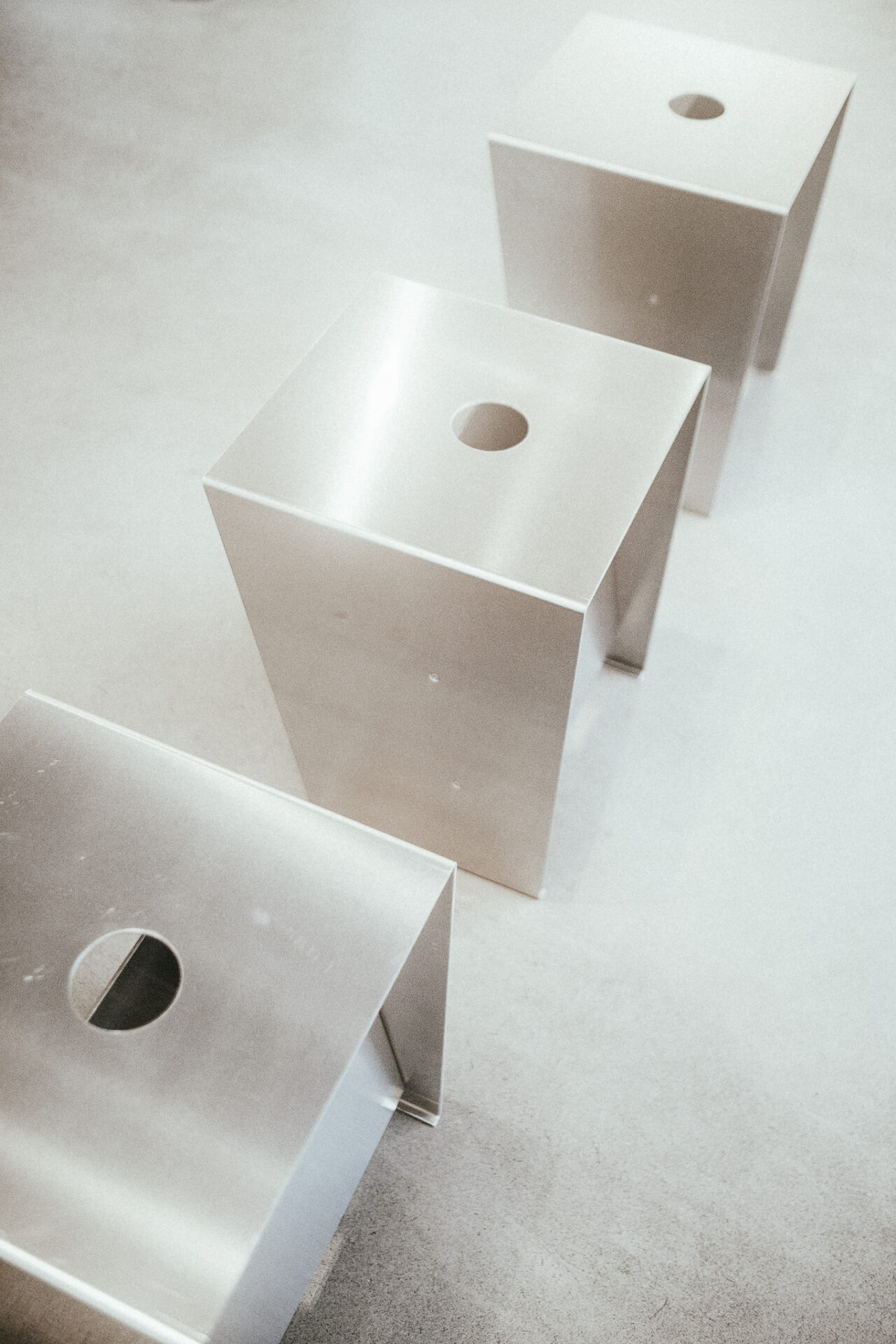 Drie doosvormige objecten van geborsteld aluminium met een rond gat in het bovenblad, schuin achter elkaar op een lichte betonvloer.